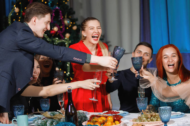 Веселе товариство друзів святкує Різдво або Новий рік за столом біля ялинки з садками. Жінки сміються, чіпляють окуляри шампанським.. - Фото, зображення