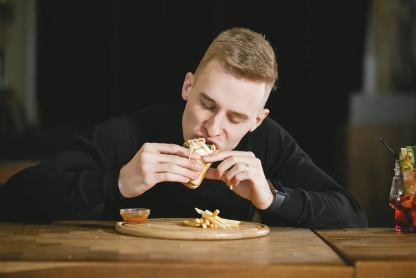 Νέος όμορφος ξανθός τύπος τρώει ένα σάντουιτς σε ένα τραπέζι σε ένα καφέ.. - Φωτογραφία, εικόνα