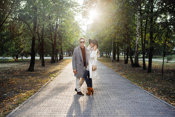 公園の舗装された路地で抱き合っているカップルを愛しています。彼は灰色のコートと黒の眼鏡で、彼女は白と帽子の中にあります。. - 写真・画像