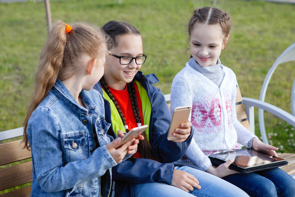 Drie tiener meisje met een smartphone zit op een bank in een zomer stadspark. - Foto, afbeelding