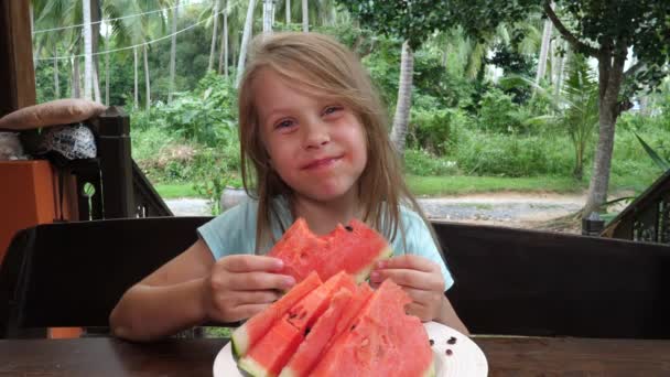 Linda niña sonriente se sienta en la mesa de madera afuera sosteniendo y mordiendo rebanada de sandía roja
 - Metraje, vídeo