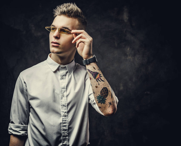 Прохолодний татуйований молодий чоловік виглядає впевнено і позує ізольовано в темній студії перед сірим тлом
 - Фото, зображення