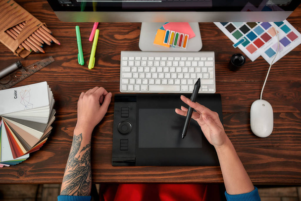 Κάνοντας δημιουργικό σχεδιασμό. Κάτοψη των αρσενικών χεριών που χρησιμοποιούν ψηφιακό tablet γραφικών και επιφάνεια εργασίας στο γραφείο ή το στούντιο. Χώρος εργασίας με γραφικά τάμπλετ, πληκτρολόγιο, υπολογιστή και δείγματα χρωμάτων - Φωτογραφία, εικόνα