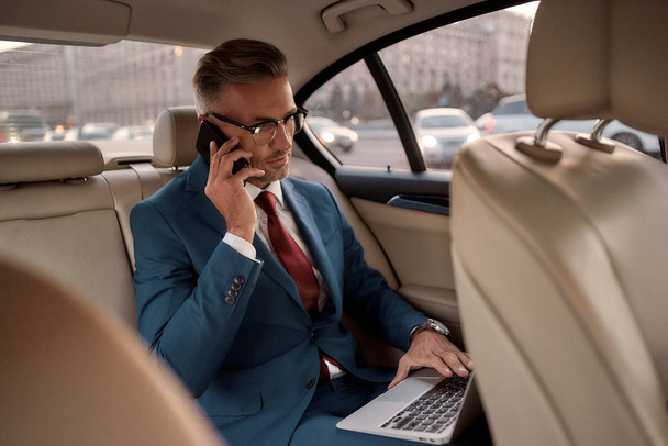 Βγάζω λεφτά. Αυτοπεποίθηση και κομψό ώριμος επιχειρηματίας σε πλήρη κοστούμι εργασίας για το laptop του και μιλώντας στο τηλέφωνο με τον επιχειρηματικό εταίρο, ενώ κάθεται στο αυτοκίνητο - Φωτογραφία, εικόνα