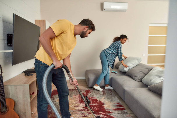 Dirty work. Happy multicultural couple in casual clothes cleaning their modern living room together. Young man cleaning the carpet with vacuum cleaner - Fotoğraf, Görsel