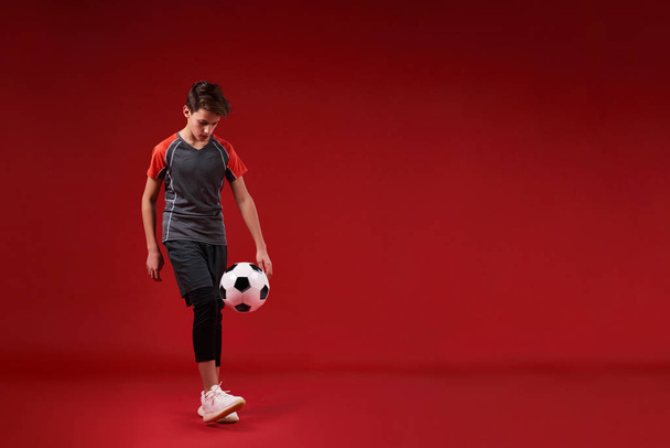 When you play ball, leave it all. A teenage boy engaged in sport, looking at football while training with it. Isolated on red background. Fitness, training, active lifestyle concept. Horizontal shot. - Photo, Image