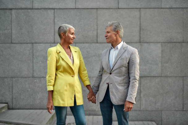Portrait of happy and beautiful middle-aged couple in stylish wear holding hands and looking at each other with smile while standing against grey wall outdoors - Valokuva, kuva