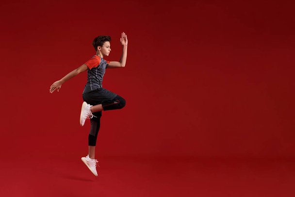 Cardio training. A teenage boy is engaged in sport, he is looking aside while jumping. Isolated on red background. Fitness, training, active lifestyle concept. Horizontal shot - Foto, Imagen