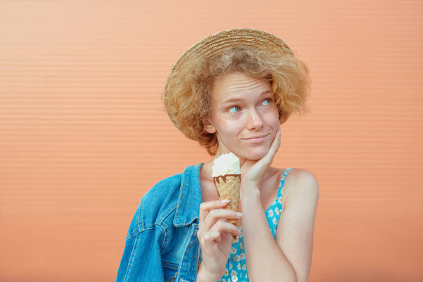 joven pelirroja rizada en sombrero de paja, vestido azul y chaqueta vaqueros comiendo helado sobre fondo beige. Diversión, verano, moda, concepto juvenil
 - Foto, Imagen
