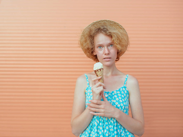 młoda, kręcona ruda kobieta w słomkowym kapeluszu i niebieskiej sukience jedząca lody na beżowym tle. Zabawa, lato, moda, koncepcja młodzieży - Zdjęcie, obraz