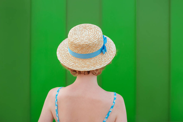plecy młodej rudej kobiety w słomkowym kapeluszu i niebieskiej sukience stojącej na zielonym tle ściany. Zabawa, lato, moda, koncepcja młodzieży - Zdjęcie, obraz