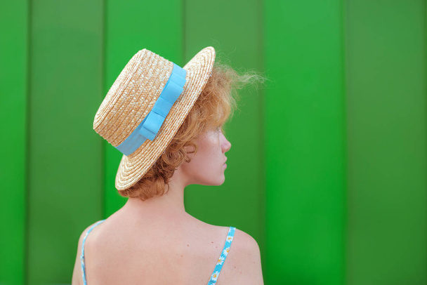 back of young curly redhead woman in straw hat and blue sundress standing on green wall background. Fun, summer, fashion, youth concept. Copy space - Zdjęcie, obraz