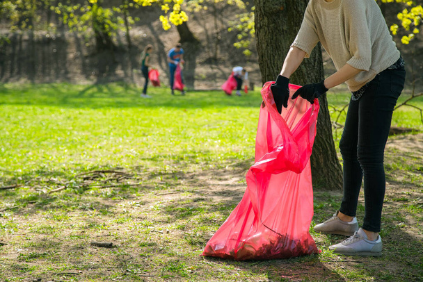 Mężczyzna i kobieta wolontariusz noszenia podnoszenia śmieci i odpadów z tworzyw sztucznych w parku publicznym. Młodzi ludzie noszą rękawice i wkładają śmieci w czerwone plastikowe torby na zewnątrz - Zdjęcie, obraz