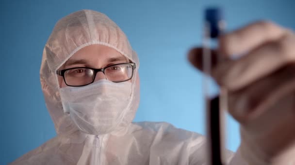 Caucasian man in white protective suit, medical mask, glasses, rubber gloves on blue background holds in hands test tube with red liquid and inscription Coronovirus - Footage, Video