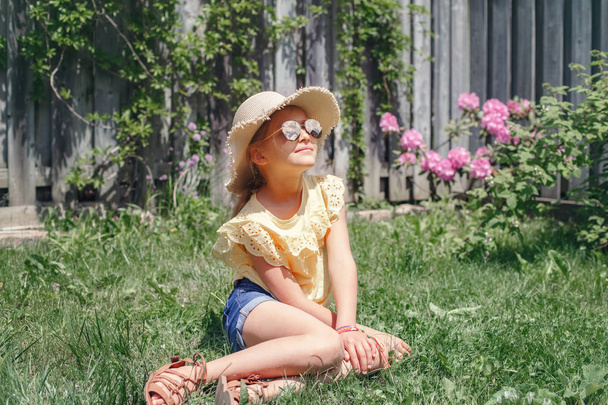 Portrait of cute adorable child girl in sunglasses and straw hat sitting on grass outdoor. Happy smiling Caucasian kid having fun at home backyard. Amazing joyful summer and lifestyle childhood. - Photo, Image