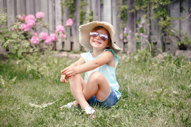 Retrato de linda niña adorable en gafas de sol y sombrero de paja sentado en la hierba al aire libre. Feliz niño caucásico sonriente divirtiéndose en el patio trasero de casa. Increíble verano alegre y estilo de vida de la infancia. - Foto, imagen