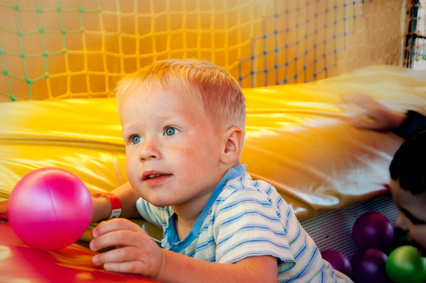 Ein blonder Junge von drei Jahren in einem Spielcenter spielt in einem Pool mit farbigen Plastikbällen - Foto, Bild