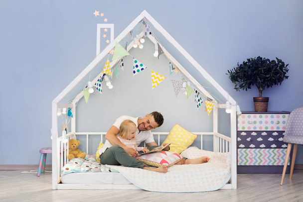 Happy father dad in cute house bed with blonde daughter reading stories at bedtime - Foto, Imagen