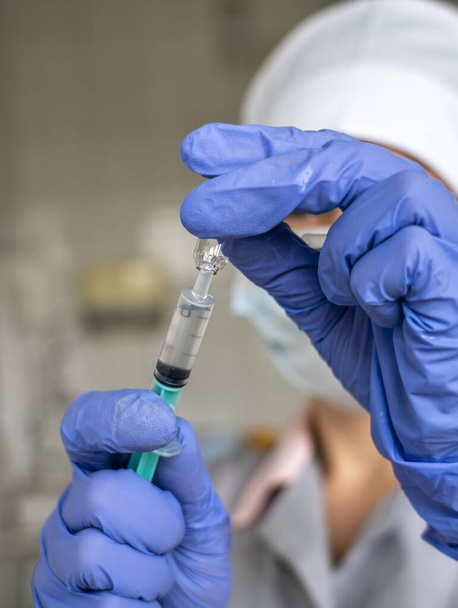 pracownik medyczny w zbliżeniu w niebieskich rękawiczkach wypełnia strzykawkę medyczną zawartością ampułki ze szczepionką. - Zdjęcie, obraz