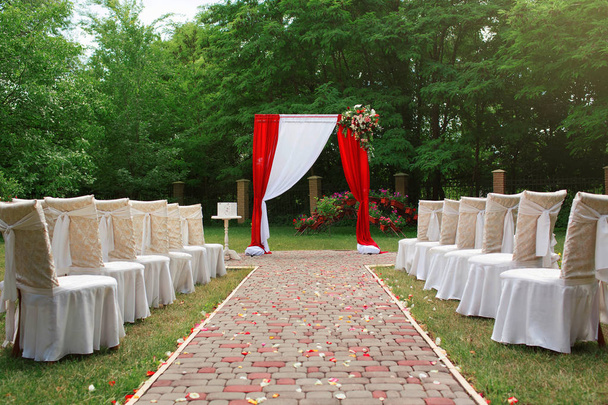 ein wunderschöner Hochzeitsbogen mit Blumen, weißen Stühlen für Gäste, Rosenblättern, einem wunderschönen Park für eine Hochzeitszeremonie, einer perfekten Hochzeit - Foto, Bild