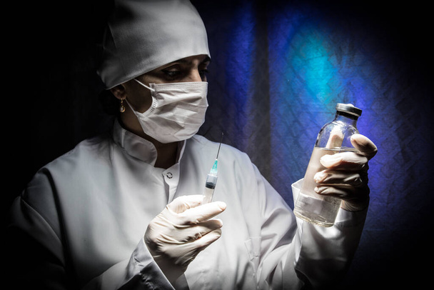 Gezondheidsconcept. Ernstige vrouwelijke arts in medisch masker met spuit. Gezondheidswerker draait het vaccin in een spuit op donkere achtergrond. - Foto, afbeelding