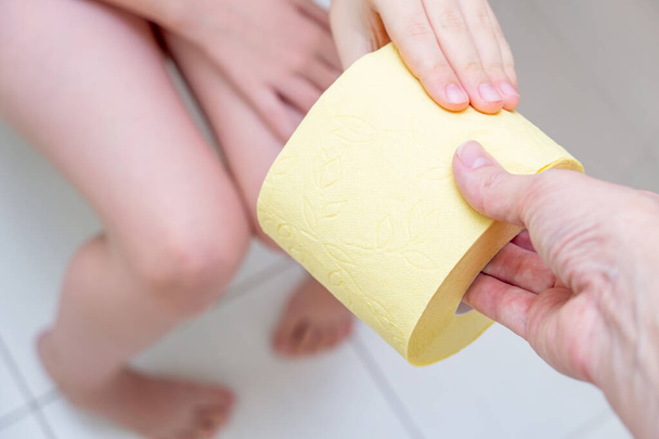 Mother giving a roll of yellow toilet paper to her son sitting on a toilet, child digestive problems and difficulties in toilet concept. - 写真・画像