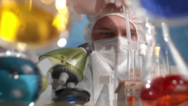 Caucasian bearded man is pipetting and mixing red liquid into beaker and onto microscope stand, shakes and mixes chemicals. On glass transparent table are vessels, flasks with multi-colored solutions - Materiaali, video