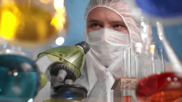 Tıbbi maske ve koruyucu tulum giyen bilim adamları pipetten şişeye portakal suyu düşürür, sallar, kimyasalları karıştırır. Cam şeffaf masa üzerinde çok renkli çözeltilere sahip kaplar ve mataralar var. - Video, Çekim