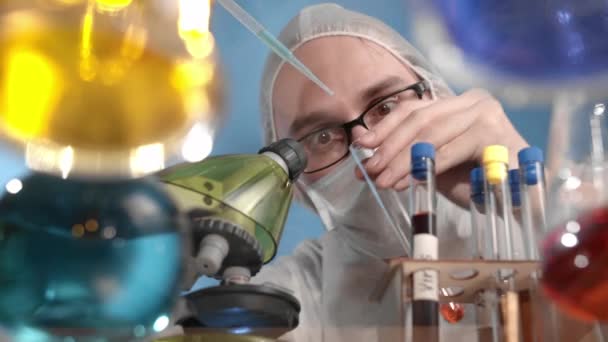 A tudósok szemüveget viselnek orvosi maszkban és védőruhát, piros folyadékot csepegtetnek pipettáról főzőpohárba. Az üveg átlátszó asztal edények és lombikok többszínű oldatok, vírus - Felvétel, videó