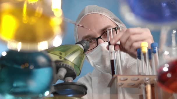 Científicos que llevan gafas en máscara médica y protectores descubren el líquido del virus rojo de la pipeta a la colmena. En la mesa transparente de cristal son los vasos y las manchas con las soluciones multicolores. - Imágenes, Vídeo