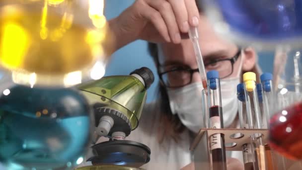 Caucasian bearded man wearing glasses is pipetting and mixing red liquid into beaker and onto microscope stand. On glass transparent table are vessels, flasks with multi-colored solutions - Materiaali, video