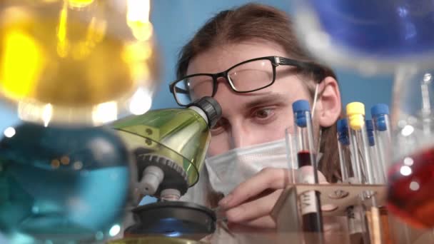 L'uomo barbuto caucasico sta pipando sul cavalletto del microscopio, si rallegra della scoperta e beve acqua arancione. Sul tavolo trasparente in vetro ci sono vasi, fiaschi con soluzioni multicolore - Filmati, video