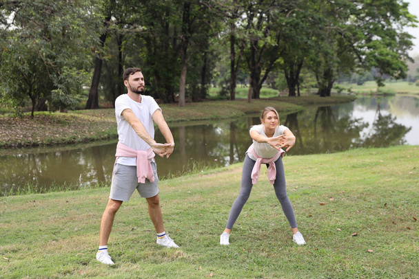 image of young caucasian couple enjoying in exercising and stretching outdoor with smiling faces and green trees during summer time in the park - Foto, Imagen