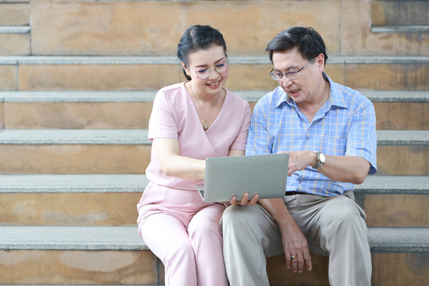 シニアアジア旅行カップル夫と妻でピンクと青のシャツに白い素敵な帽子が座っていますダウンタウンの階段に座って、休憩を取りながら場所を検索するためにコンピュータを使用して.  - 写真・画像