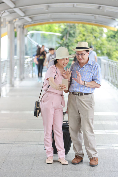 フル長さアジア旅行カップル夫と妻ピンクと青のシャツで白い素敵な帽子と荷物が幸せな笑顔でダウンタウンを歩くと退職年齢の間に場所を検索するためにモバイルを使用して - 写真・画像