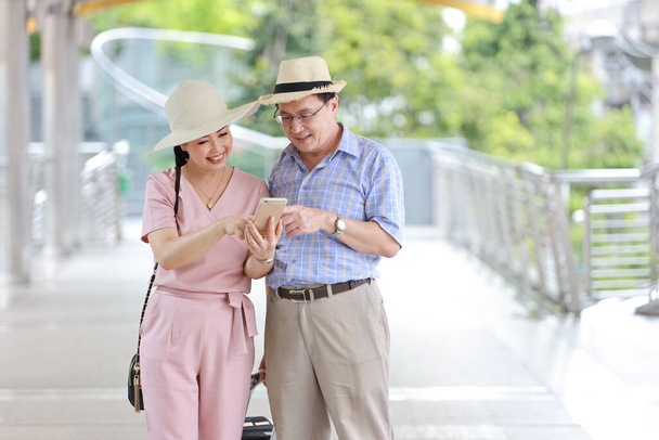 idős ázsiai utazó pár férj és feleség rózsaszín és kék ing fehér szép kalap és poggyász séta a belvárosban boldog mosolygós arc és a mobil keresési hely nyugdíjas korban - Fotó, kép