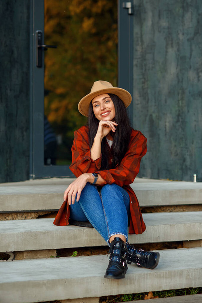 Beautiful woman in a red shirt, jeans and hat sits on the stairs of her modern home near the forest. - Foto, afbeelding