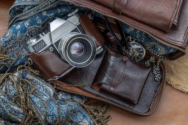 Top view of a vintage photo camera and a brown leather bag with scarf, glasses and pocket watch on sack cloth background. Holiday traveling concept design. Vintage color tone. - Photo, image