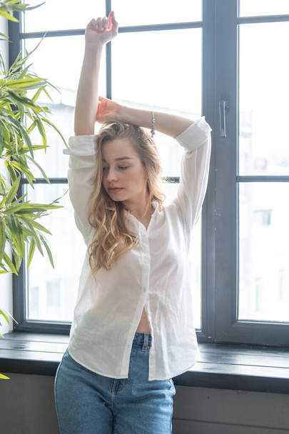 Young beutiful blond woman wearing white shirt and blue jeans standing next to window. Morning portrait of a girl with window and green plants on the background - Photo, image