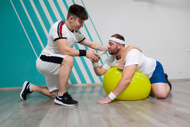 Overweight man is lying on the fitness ball in the gym while his personal trainer offers him a bottle of water. Fat man is dying of thirst and looking exhausted after a hard training in fitness group - 写真・画像