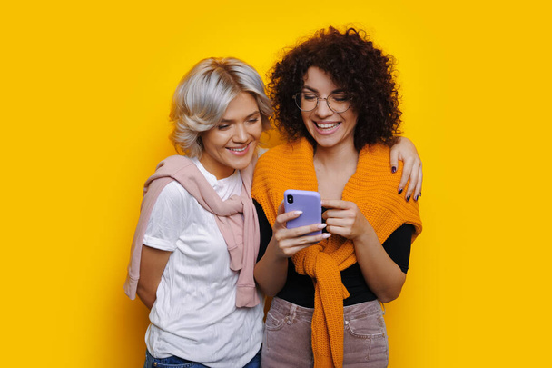 Urocza biała dziewczyna z kręconymi włosami i okularami pokazuje coś przez telefon swojemu przyjacielowi podczas pozowania na żółtej ścianie - Zdjęcie, obraz