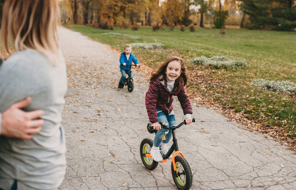 Bambini caucasici in sella alle loro biciclette e sorridenti felicemente ai loro genitori durante una passeggiata insieme nel parco
 - Foto, immagini