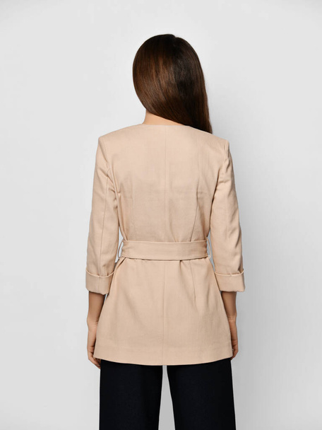 Young brunette woman is posing backwards in stylish beige formal suit jacket with cloth belt - Zdjęcie, obraz