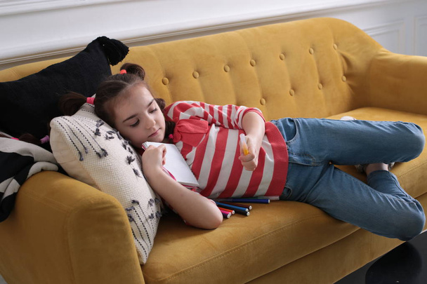 Une fillette de huit ans de bonne humeur en chandail rayé avec une coiffure drôle s'assoit sur un canapé brun avec un bloc-notes et un crayon feutre entre les mains - Photo, image