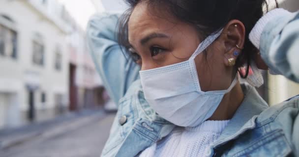 Side view close up of a mixed race woman with dark hair out and about in the city streets during the day, wearing a face mask against air pollution and coronavirus, standing and fastening her face mask with buildings in the background in slow motion. - Záběry, video