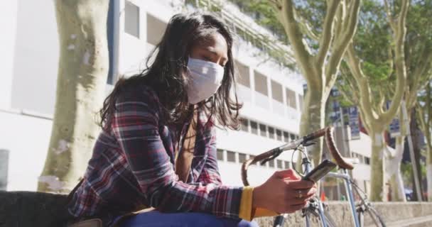  mixed race woman with dark hair out and about in the city streets during the day, wearing a face mask against air pollution and coronavirus, sitting on steps using a smartphone, a bike next to her with buildings in the background in slow motion - Imágenes, Vídeo