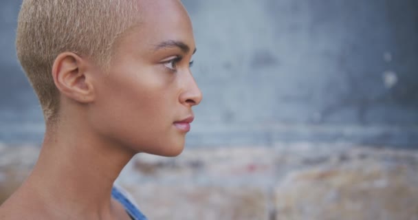Portrait close up of happy mixed race woman with short dyed blonde hair out and about in the city on a sunny day, smiling and standing against the wall in slow motion. - Кадры, видео