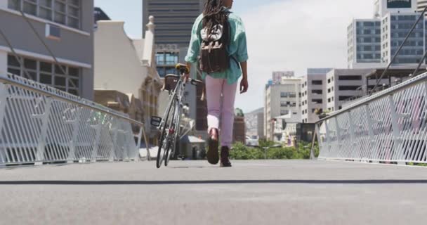 Rear view of a mixed race man with long dreadlocks out and about in the city on a sunny day, wearing backpack, walking the street and wheeling his bicycle in slow motion. - Imágenes, Vídeo
