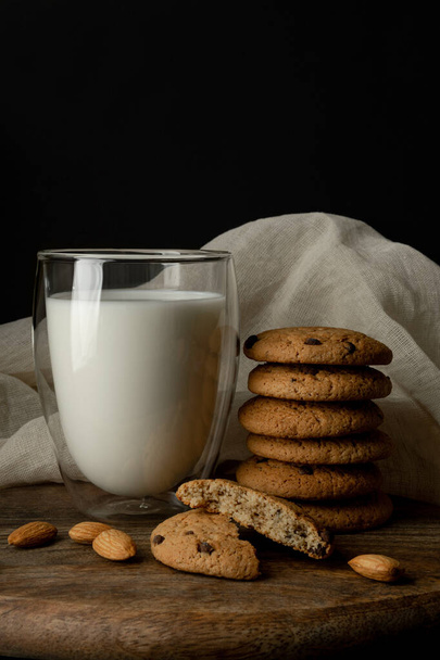 Oat cookies with pieces of chocolate, almond. Milk in a glass with double walls. One cookie is broken in half. On cutting board and white textile - Foto, afbeelding