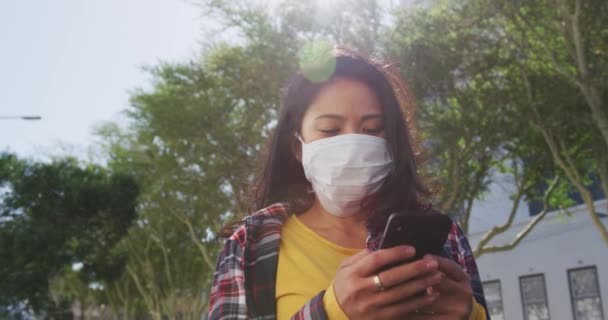  mixed race woman with long dark hair out and about in the city streets during the day, wearing a face mask against air pollution and coronavirus, walking in a city street and using a smartphone with buildings in the background in slow motion. - Záběry, video